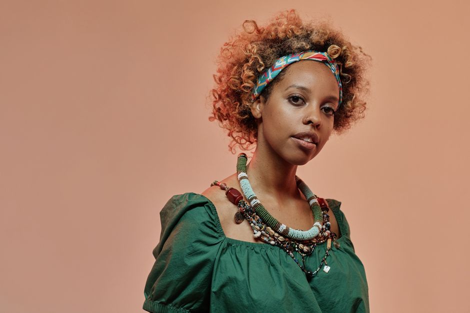 A beautiful woman with afro hair and stylish necklaces poses against a warm brown background.