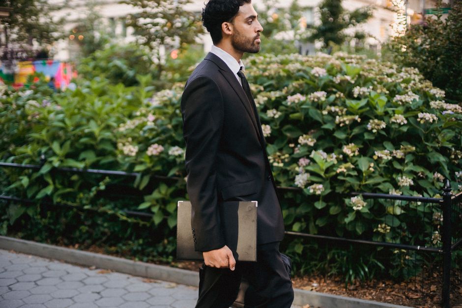 Side view of a well-dressed man walking through a city park holding a laptop.