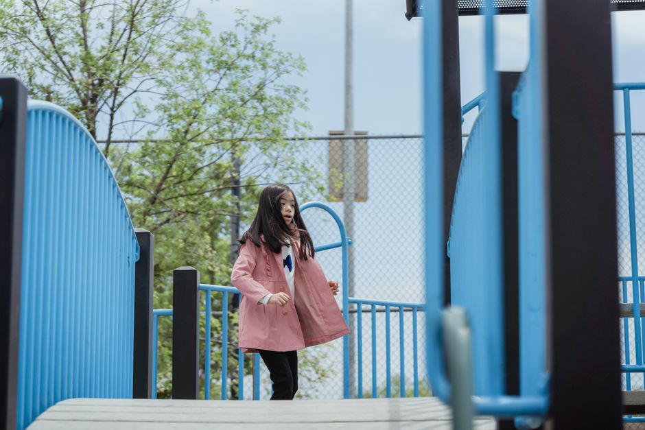 A child in a pink jacket enjoys a playground with metal railings outside.