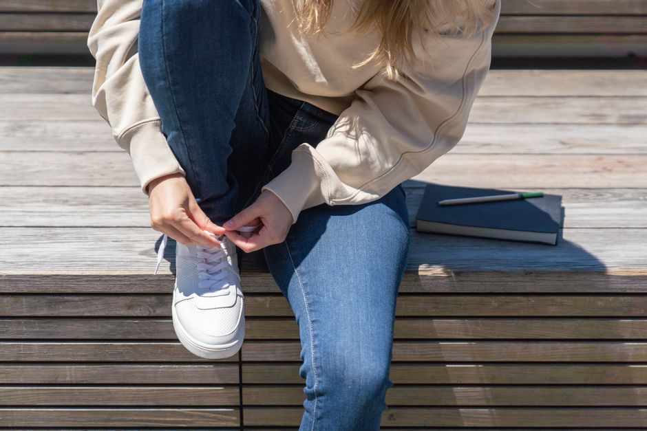 A woman in denim tying her white sneakers while sitting on outdoor wooden steps next to a 