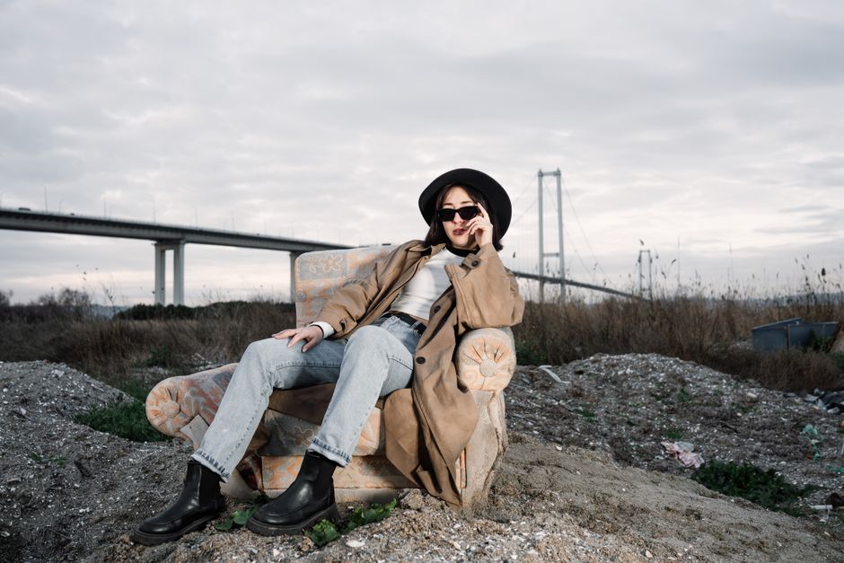 Stylish woman in a trench coat and hat lounging outdoors near a bridge, exuding a modern vibe.