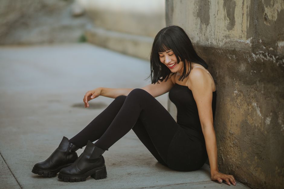A relaxed woman smiling while sitting against an urban wall in black attire.