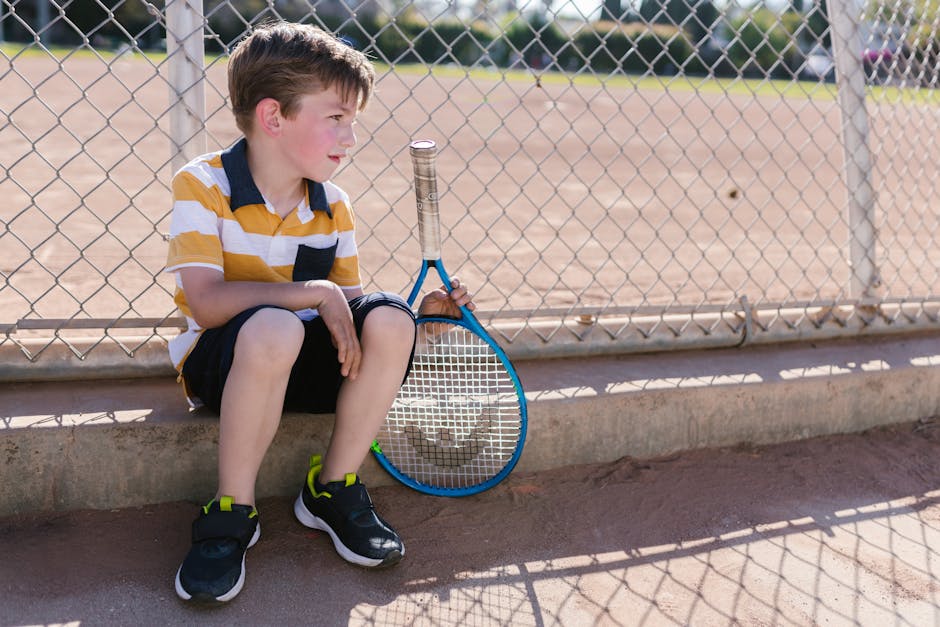A young boy sits with a tennis racket by a chain-link fence on a sunny day, embodying youthful leisure.
