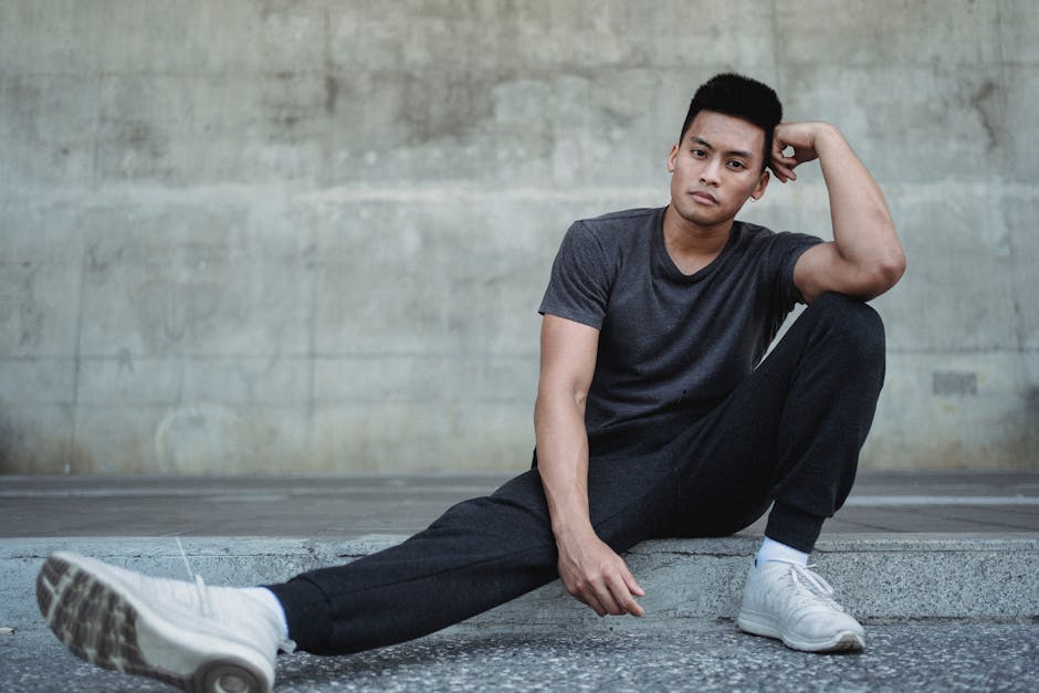 Full body of serious Asian male wearing sneakers and sportswear sitting on pavement and leaning head on hand and spread legs while looking at camera