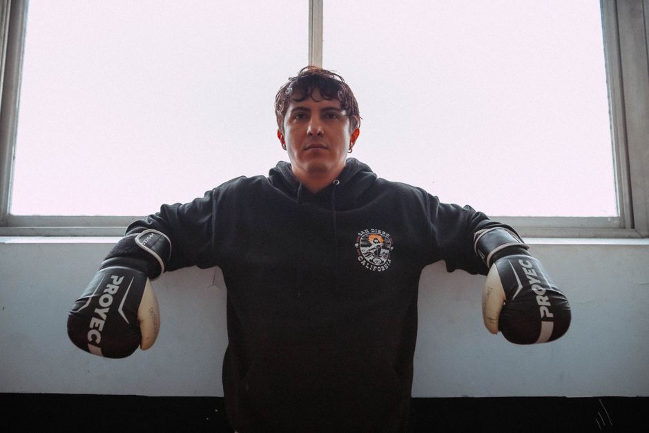A male boxer wearing gloves leans against a gym window, exuding determination and focus.
