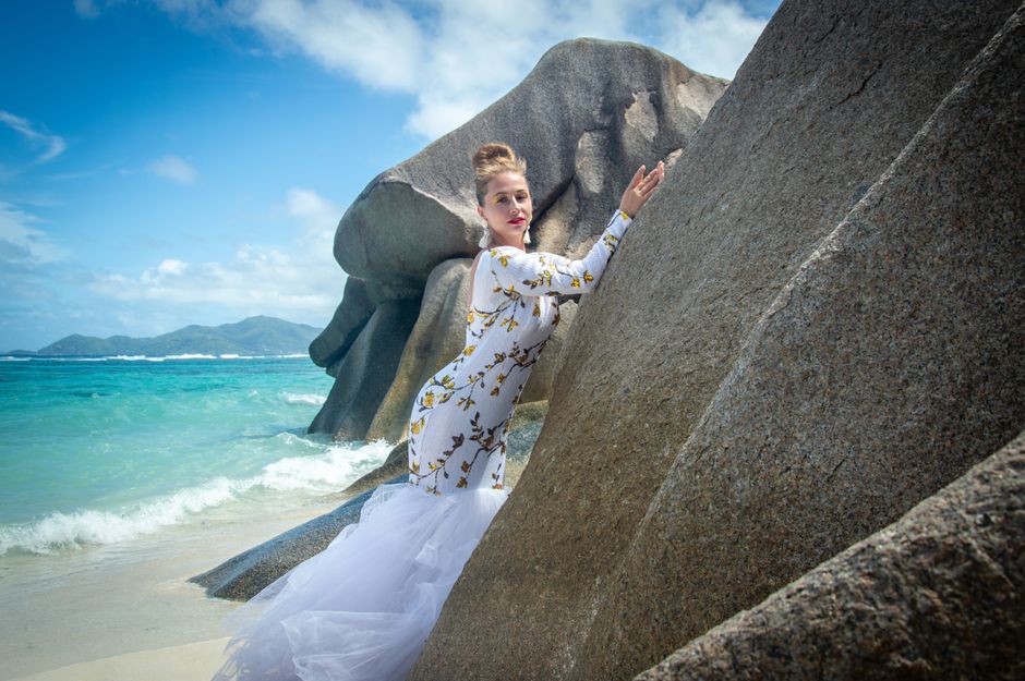 A woman in a floral dress poses dramatically against granite rocks by the sea on a sunny day.