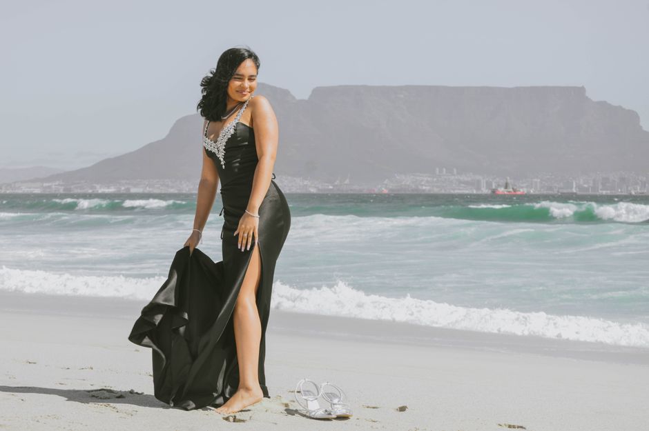 A woman in a black elegant dress poses on a beach with Table Mountain in the background.