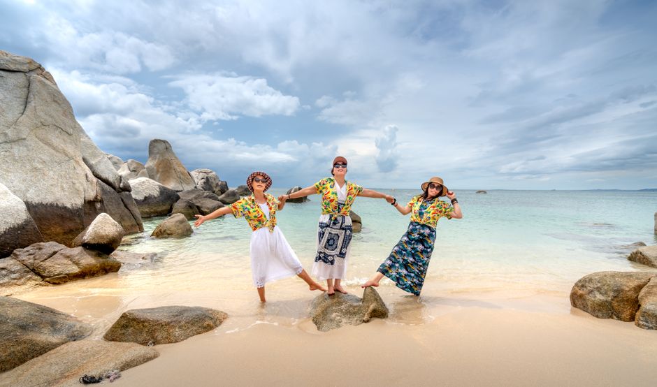 Three women enjoying a sunny tropical beach, posing on rocks with vibrant yellow shirts.