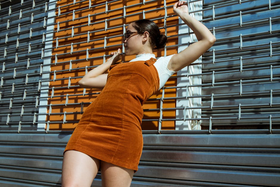 Stylish woman in orange corduroy dress and sunglasses leaning on a metal gate, striking a pose.