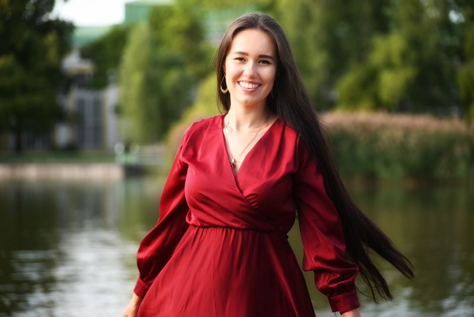 A cheerful woman in a red dress smiling by a serene lakeside on a sunny day.