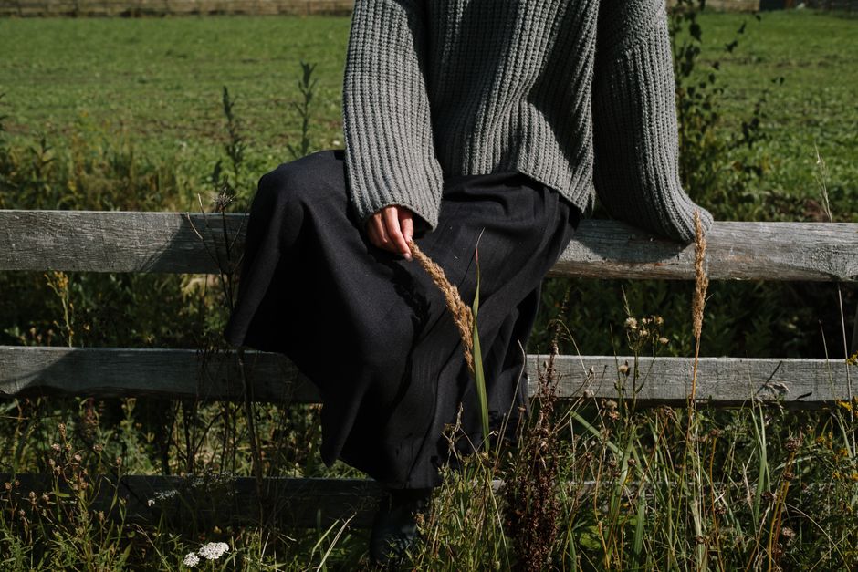 A person in a gray sweater sits on a wooden fence in a grassy rural area, creating a peaceful outdoor scene.