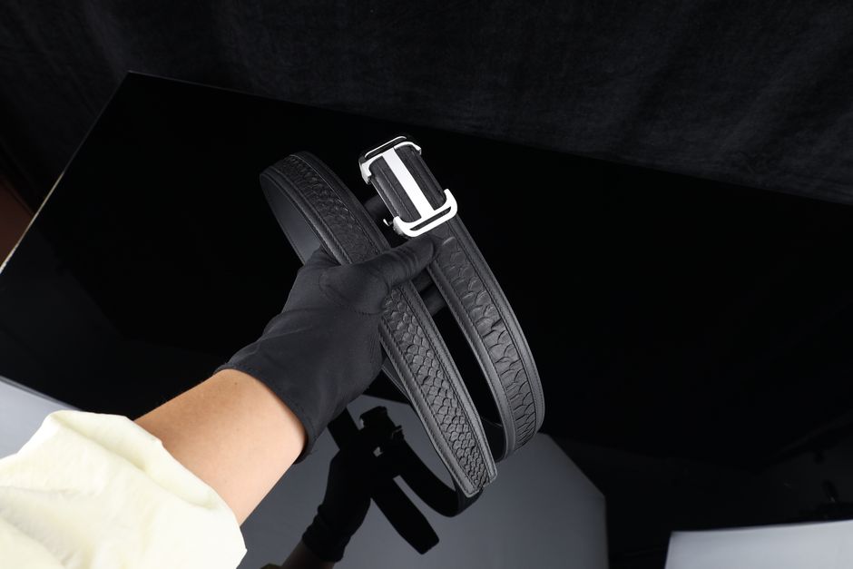 Close-up of a gloved hand holding a luxury black leather belt on a black background.
