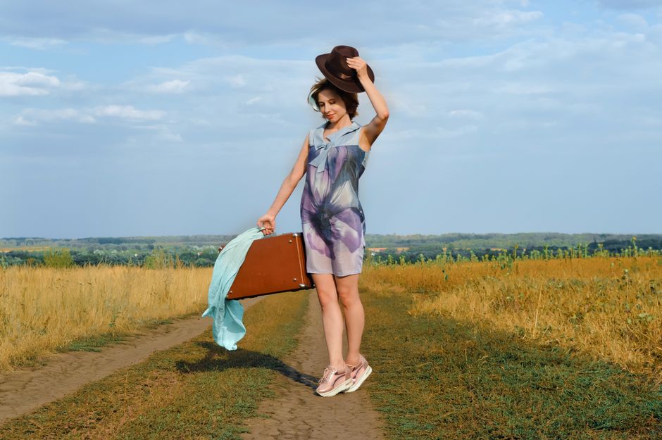 Full body young female in colorful outfit with elegant hat and vintage suitcase standing on road near fields under cloudy blue sky in countryside