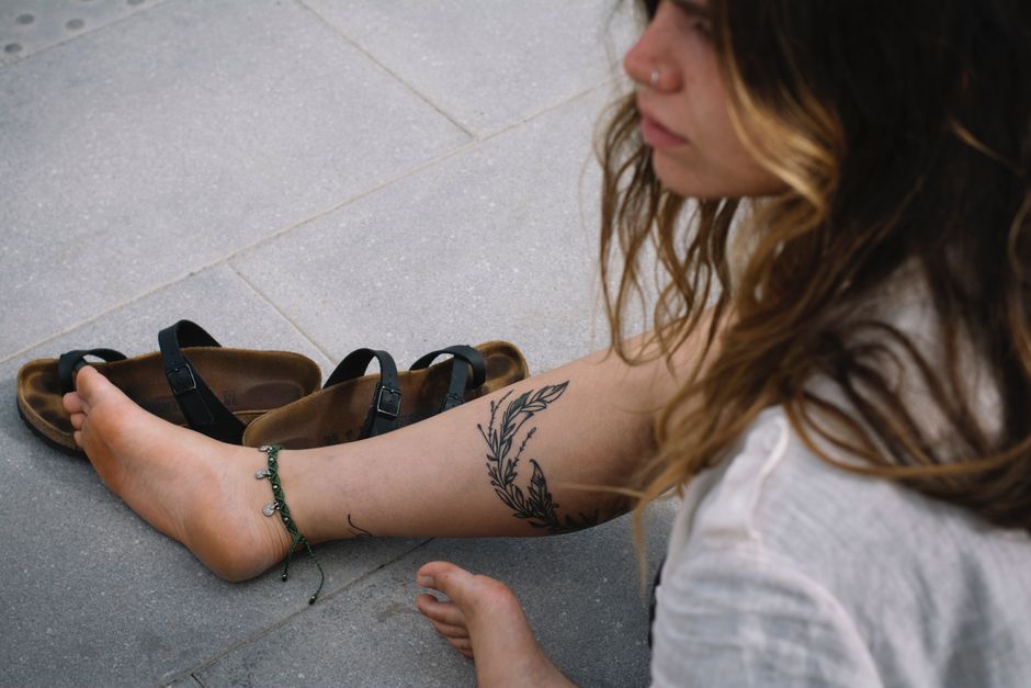 A woman sitting barefoot with a leg tattoo, enjoying a summer day on the pavement.