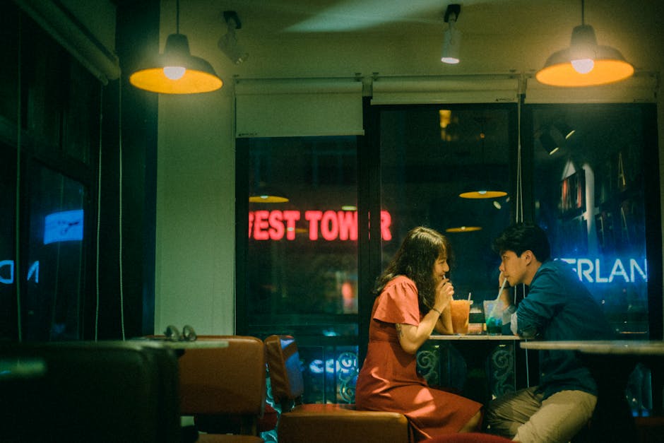 Side view of smiling ethnic woman and boyfriend drinking refreshing beverages through straws while sitting at cafeteria table with shining ceiling lamps and looking at each other in evening