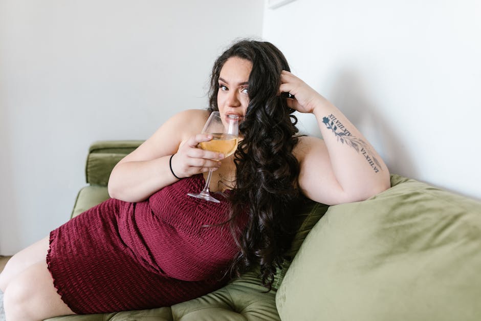 Curvy woman with arm tattoo enjoying a glass of wine while lounging on a couch, embracing body positivity.