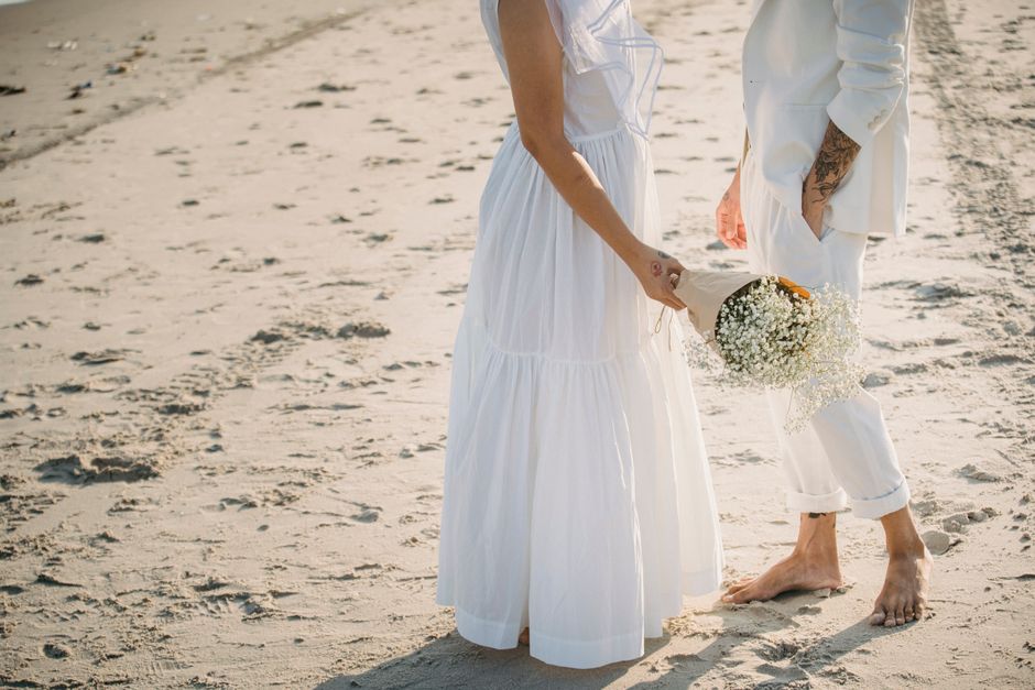 Elegant couple in white attire holding bouquet on a sunny beach.