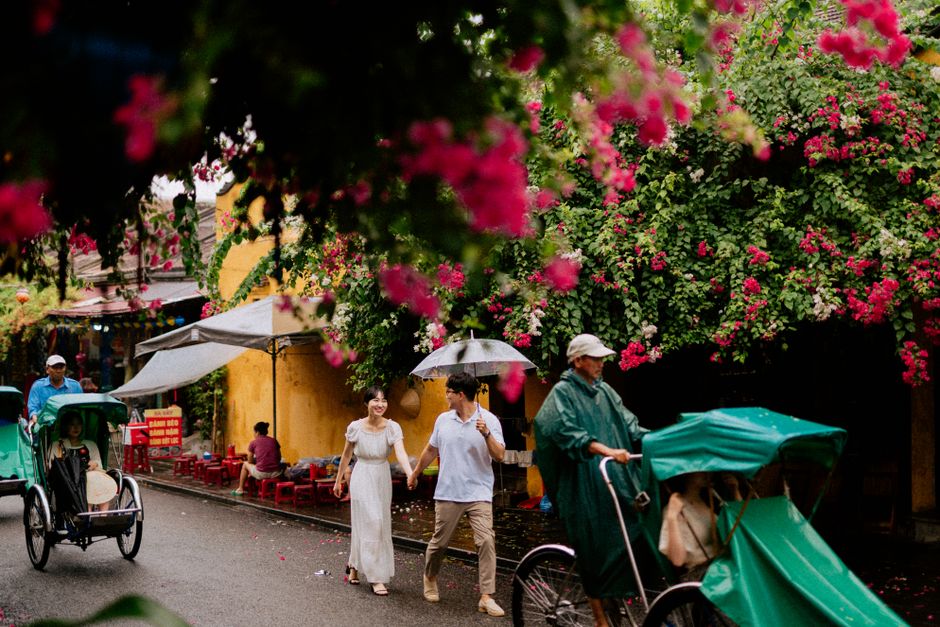 Couple walks under an umbrella in Hội An, Vietnam with vibrant blossoms overhead.