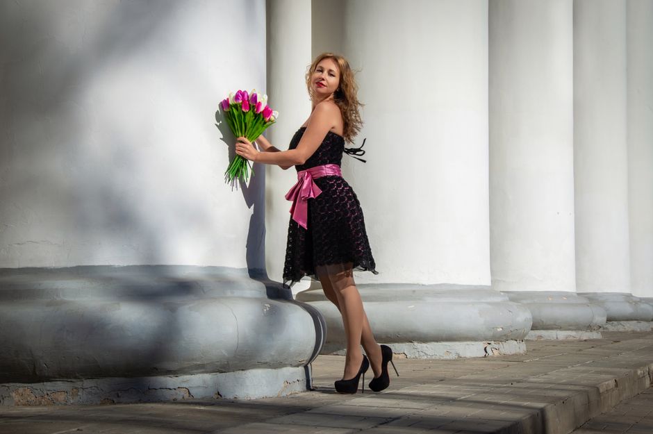 Elegant woman in a black dress with pink tulips poses against large white columns outdoors.
