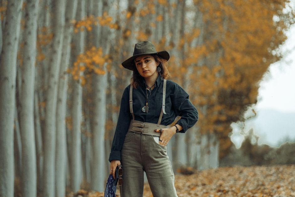 Stylish woman wearing hat and suspenders stands in autumn forest, exuding elegance.