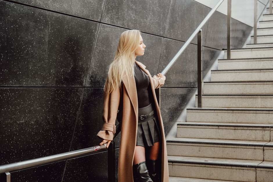 Fashionable woman in trench coat and leather boots standing by a modern staircase.