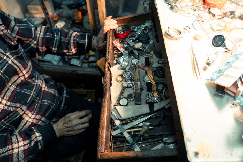 An artisan's hand at a workshop opens a drawer filled with watches, showcasing detailed repair tools.