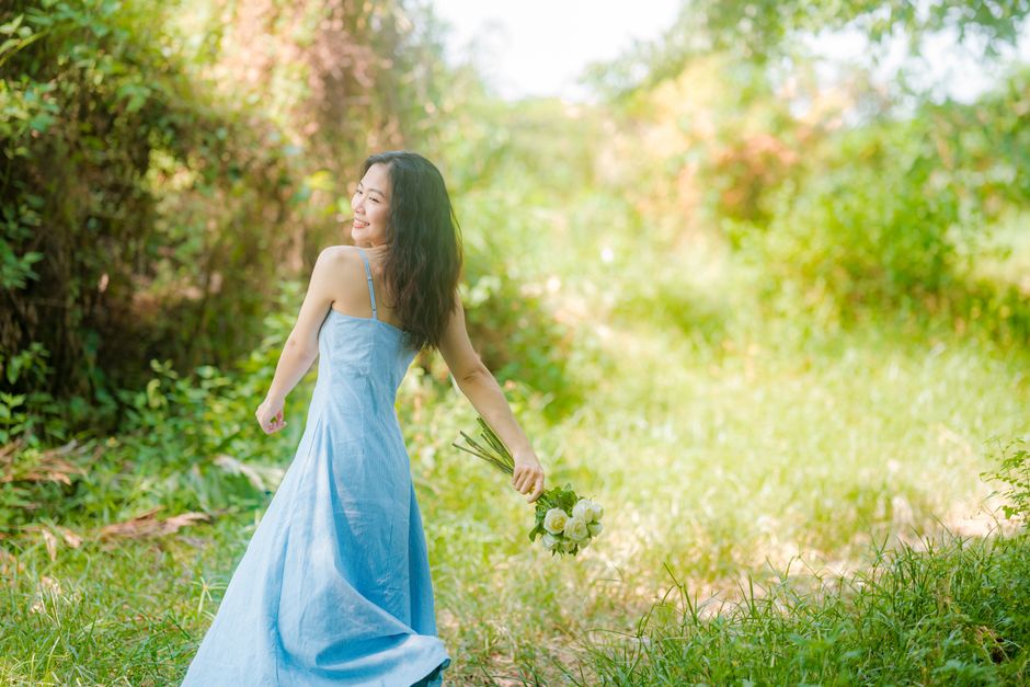 An Asian woman with long black hair in a flowing blue dress, holding flowers in a meadow.