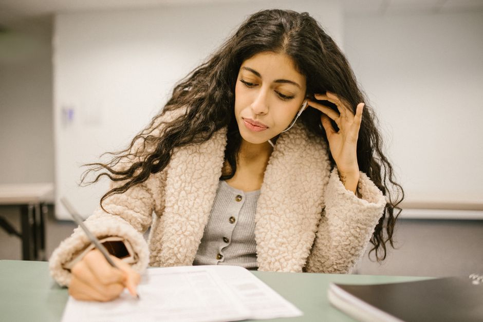 Female student writing in exam, using earphones for better focus in an educational setting.
