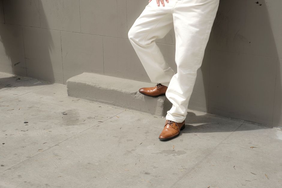 A person wearing classic brown Oxfords and white pants stands stylishly against a concrete urban wall.