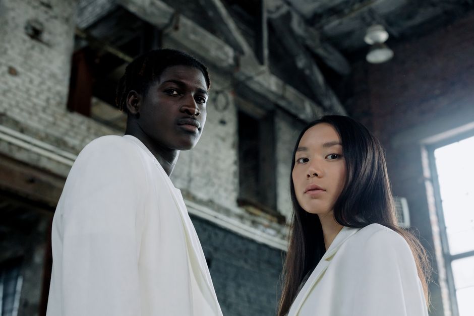 Stylish portrait of a diverse young couple in white suits, standing indoors with architectural background.