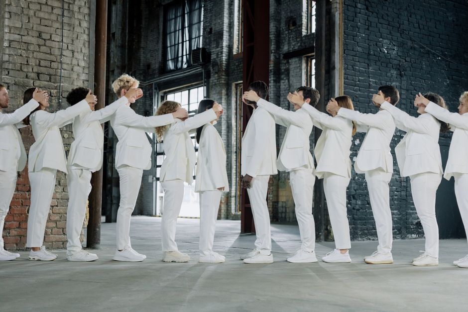 Diverse group of adults in white suits standing in a line, covering each other's eyes in an industrial setting.