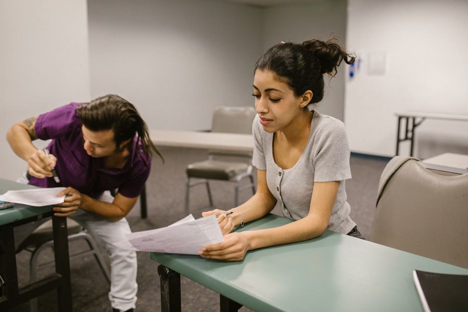 Two students in a classroom setting during an exam, one appearing to assist the other.
