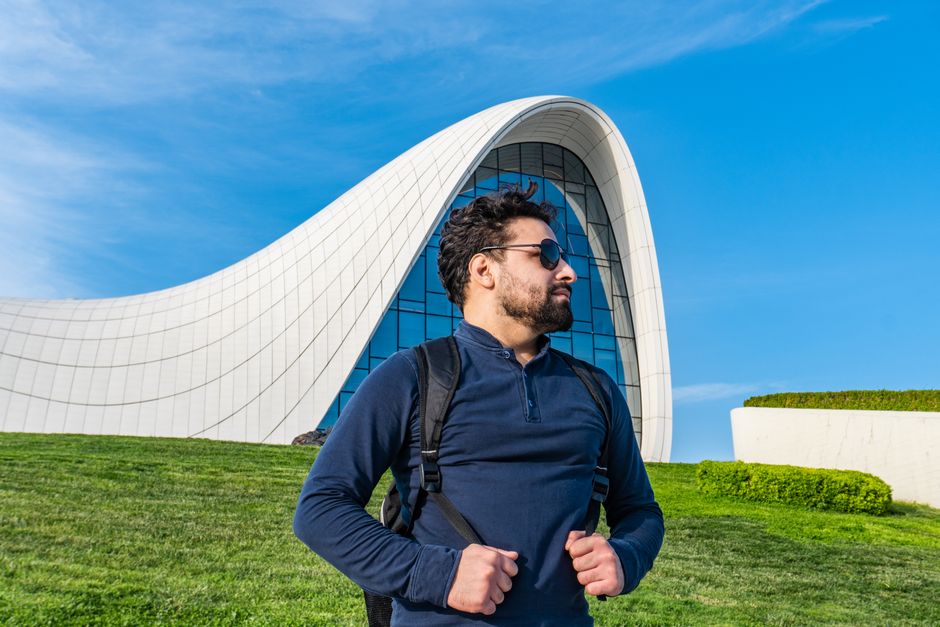 Man with backpack standing in front of modern building with curved architecture under a clear blue sky.