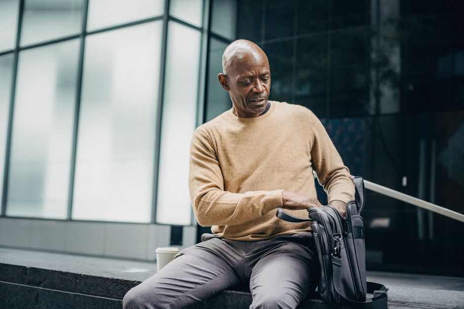 Adult bald black man in elegant jersey sitting on street bench with coffee cup and looking into bag
