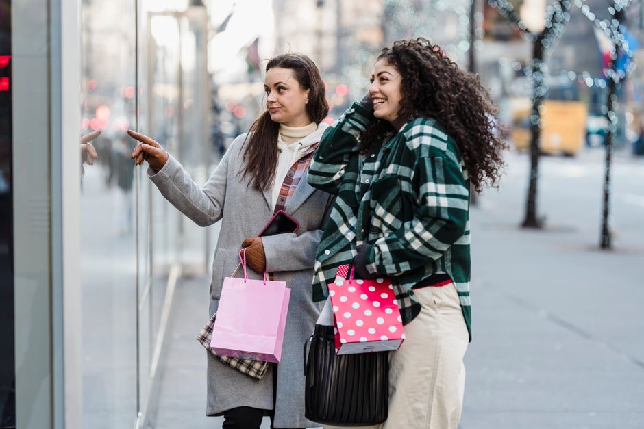 Side view of cheerful multiracial female friends with shopping bags standing on sidewalk near showcase of store on street on blurred background