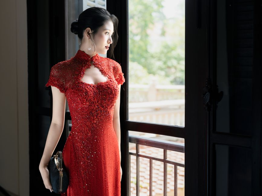 Elegant woman in red evening dress gazing outside, standing indoors.