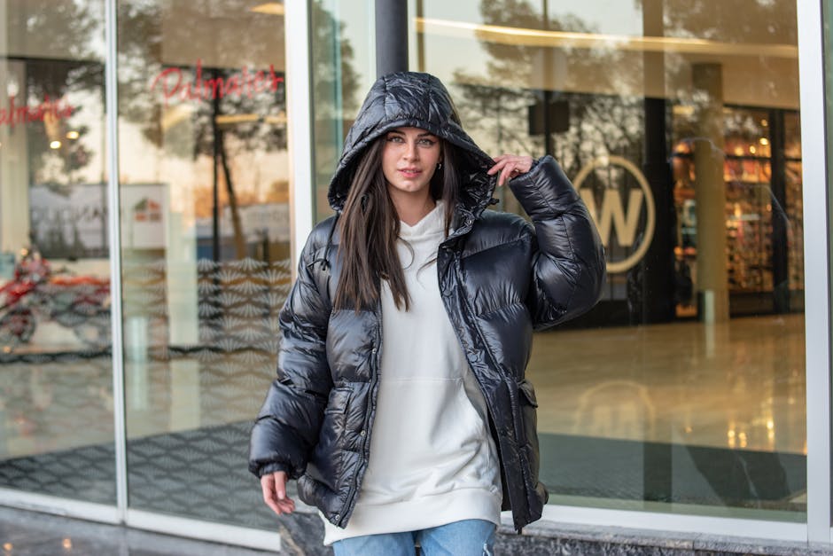 Fashionable woman with long brown hair poses in a black puffer jacket outside a store.