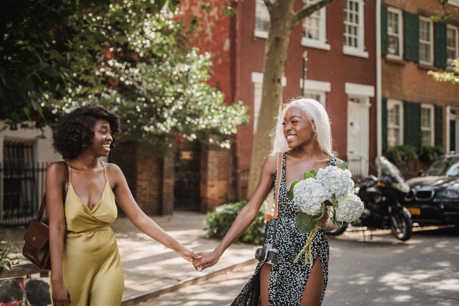 Two friends enjoy a sunny walk, holding hands and smiling, in an urban setting.