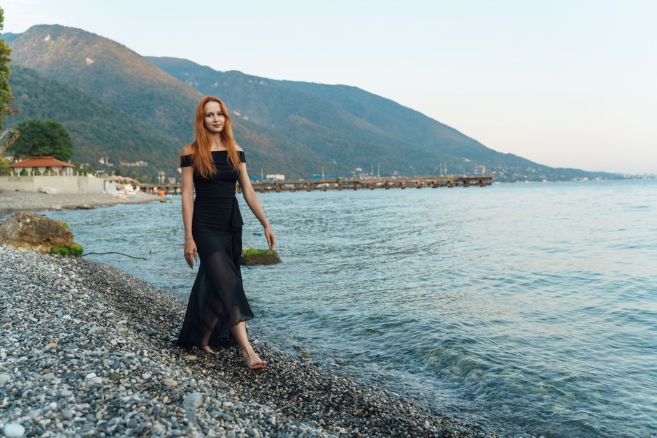 A woman in a black dress walks on a rocky beach in Гагра, with mountains and a pier in the background.