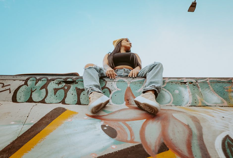 Low-angle shot of a woman sitting on a graffiti-covered wall in an urban setting, wearing casual fashion.