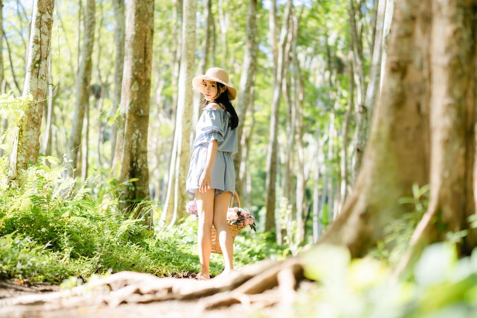 A woman in a sun hat walks through a bright forest, carrying a basket of flowers.