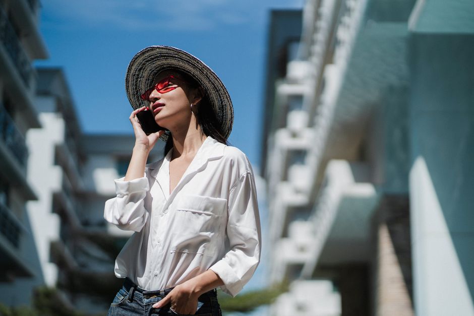 Low angle of self esteem young Asian female in stylish sunglasses and straw hat standing near modern building with hand in pocket and talking on mobile phone