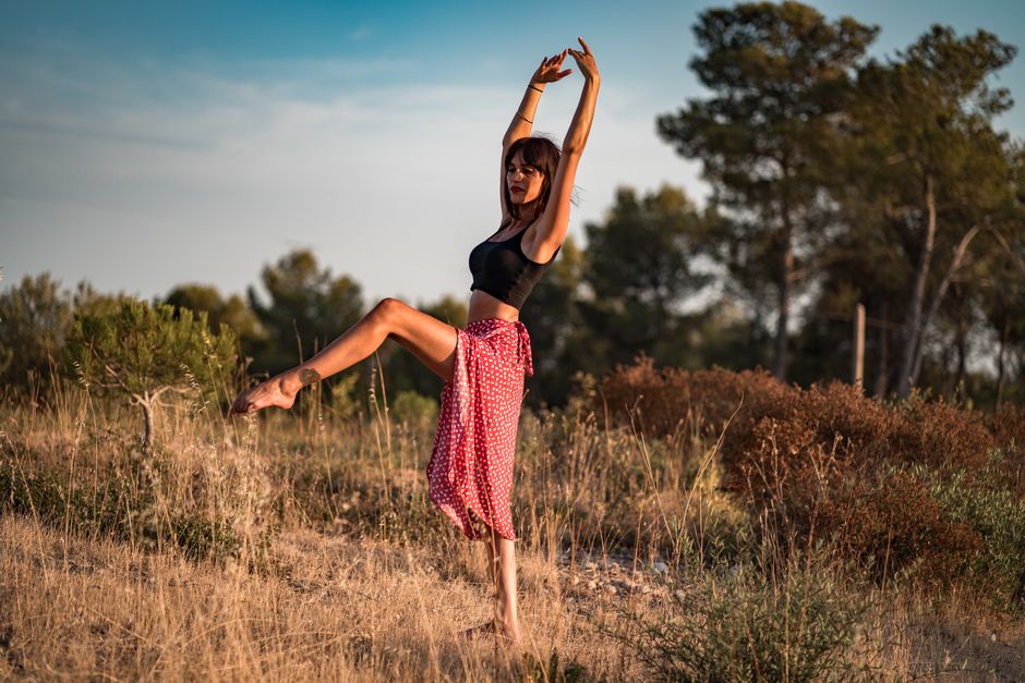 Tranquil barefoot woman in sports bra and skirt dancing with raised hands on hill in nature in sunlight