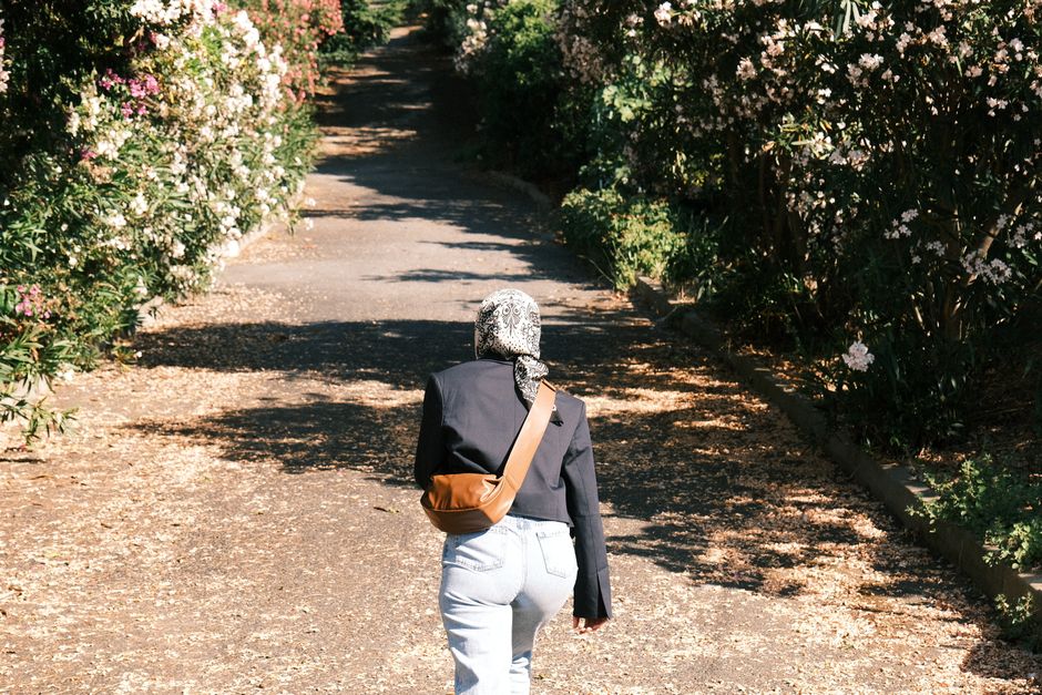 A woman walks down a sunny, tree-lined pathway, surrounded by blooming flowers and greenery.