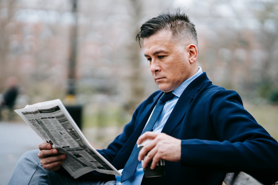 Professionally dressed man reading a newspaper on a park bench, sipping coffee.