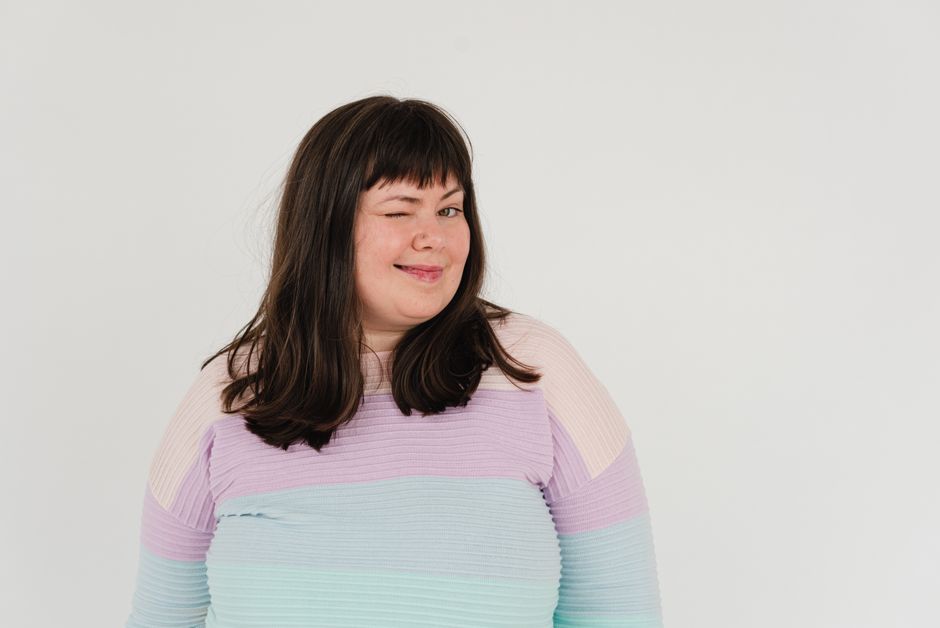 Positive plus size female in colorful sweater and dark hair looking at camera while winking on white background in light studio