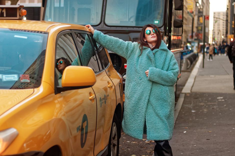 Stylishly dressed woman in a blue coat poses with a taxi on bustling city street.