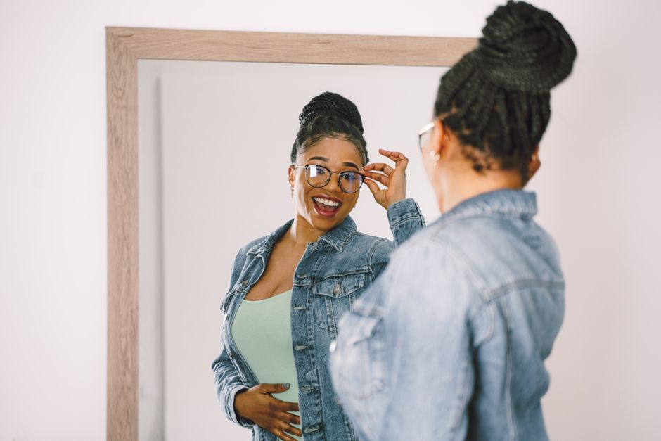 Cheerful woman in denim and glasses smiles at her reflection in a mirror.