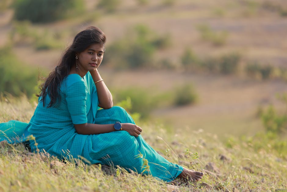 Young woman in a blue dress sits thoughtfully on a grassy hillside.