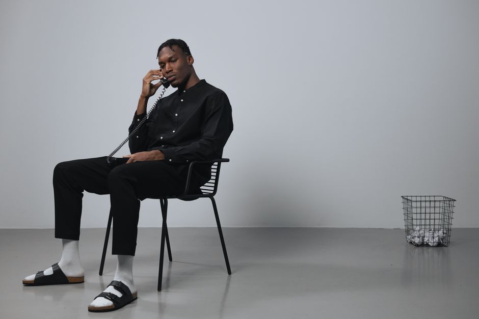African American man in black attire talking on a phone while sitting indoors with a minimalistic setup.
